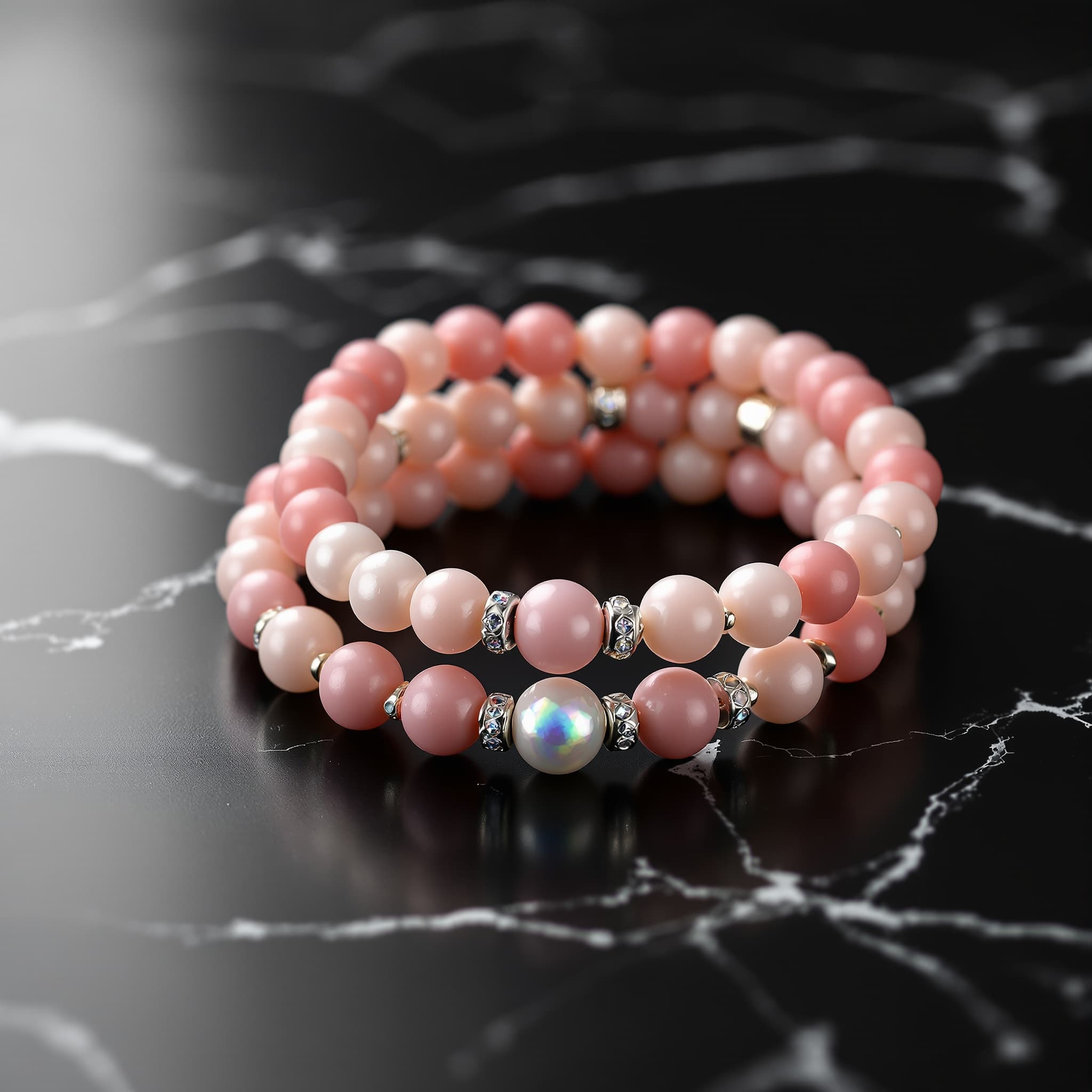 Macro product photography of a stacked set of handmade beaded bracelets, matte frosted pink beads, white pearl beads, iridescent AB crystal disco ball accent bead, silver rondelle spacers, draped on a smooth black marble surface, single soft directional studio light from upper left, deep shadows, luxury jewelry photography, minimal composition, extreme detail on bead texture, dark elegant background, no people, editorial product shot