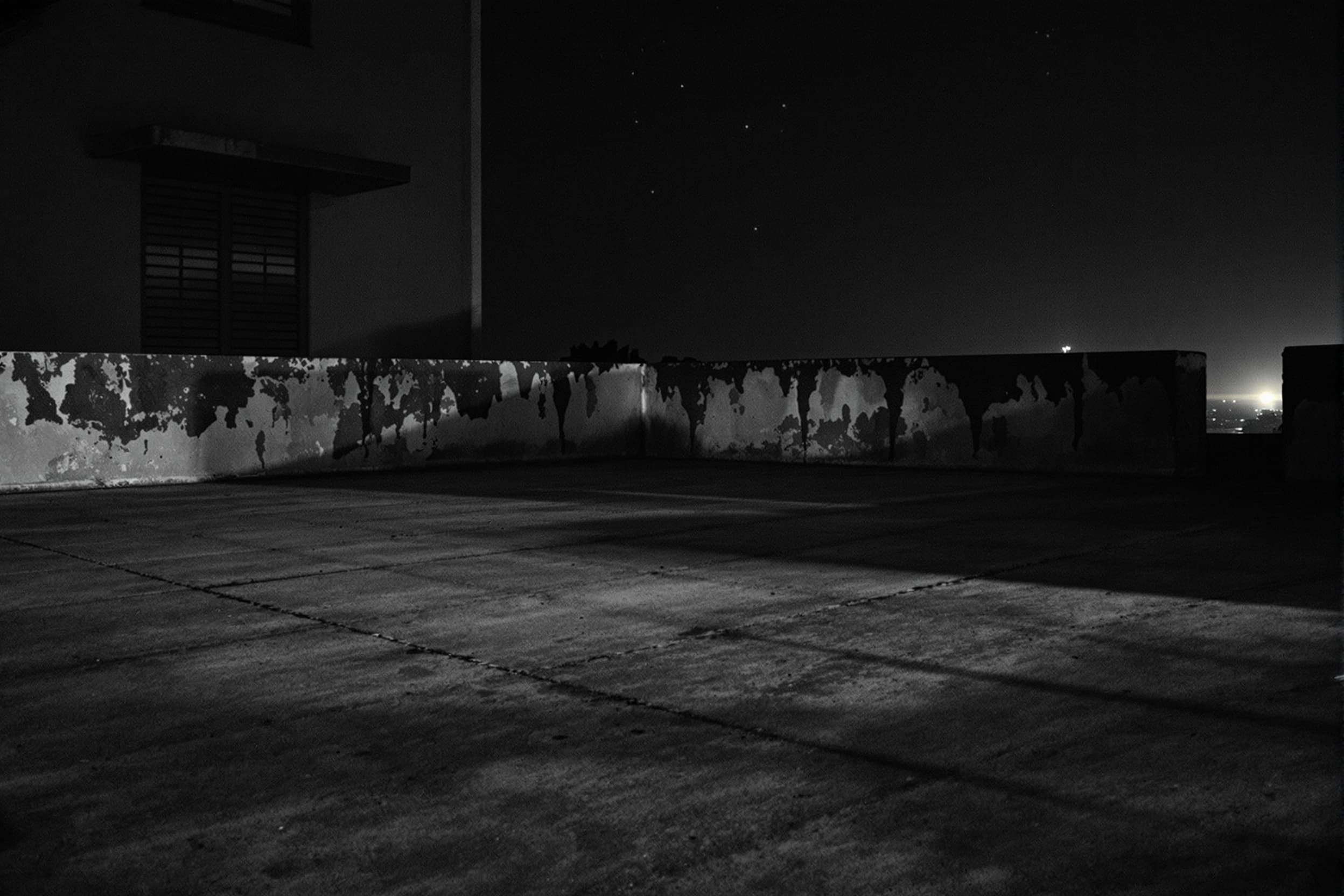 Create an archival image of a Chengalpattu in Tamil Nadu, India, empty concrete rooftop terrace surface at night, looking across the textured floor towards the weathered concrete parapet wall and the dark shuttered windows of a neighbouring building, subtle haunting darkness with long stark shadows, unsettling isolation and heavy silence, realistic and restrained dim ambient starlight and a soft distant yellow glow with a grainy 1990s archival photo texture, low angle shot from surface level --ar 16:9